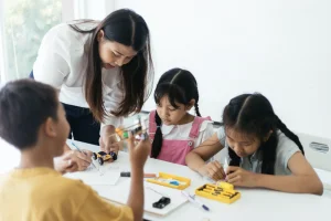 Teacher helps three children with a drawing activity at a white table, with markers and paper nearby.