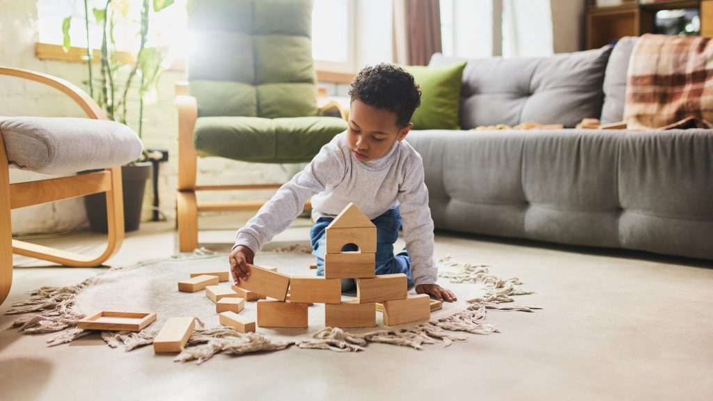 Child kneeling on a cream rug, stacking wooden blocks to build a small tower in a sunlit living room.