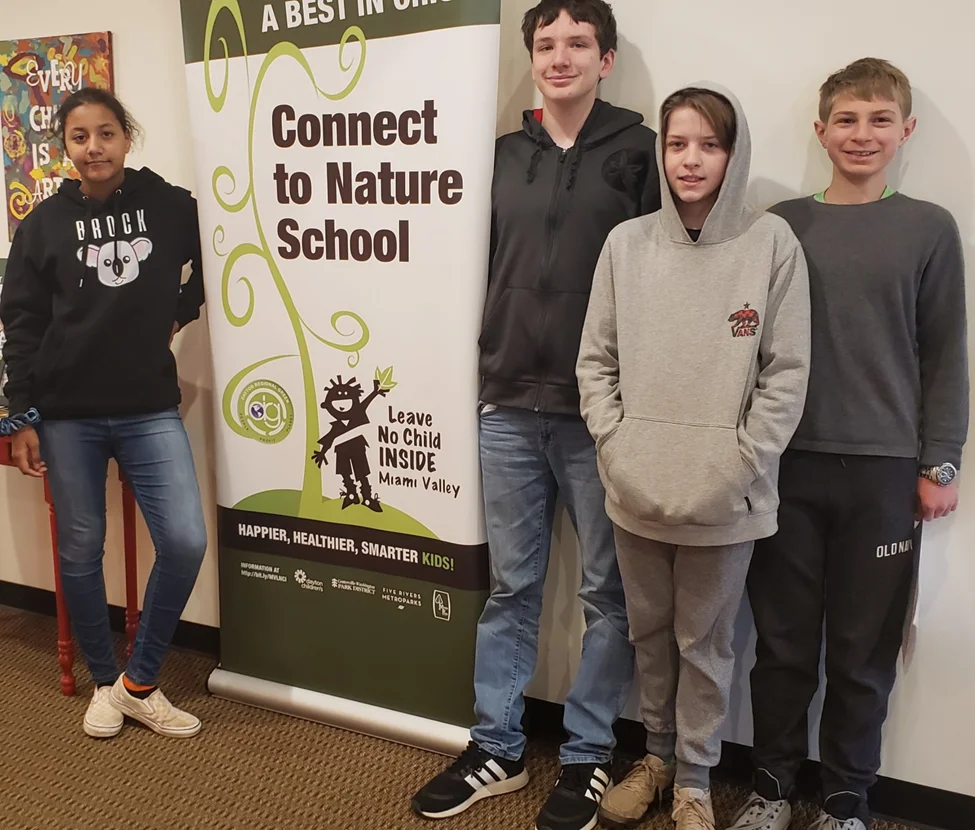 Four youths pose indoors beside a 'Connect to Nature School' banner at a Leave No Child Inside event.
