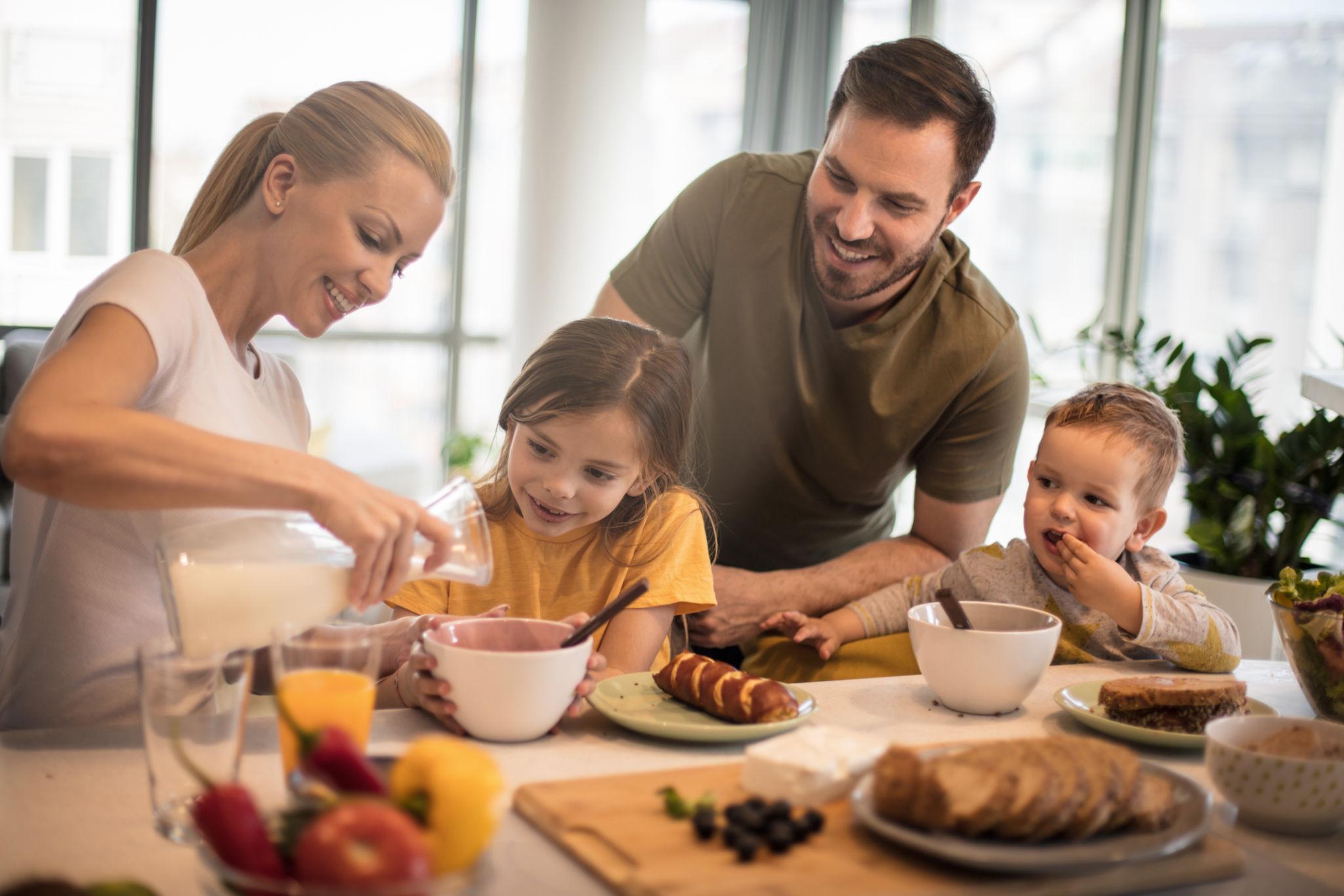 A smiling family of four enjoys breakfast together at a bright kitchen table with bowls of cereal and pastries.