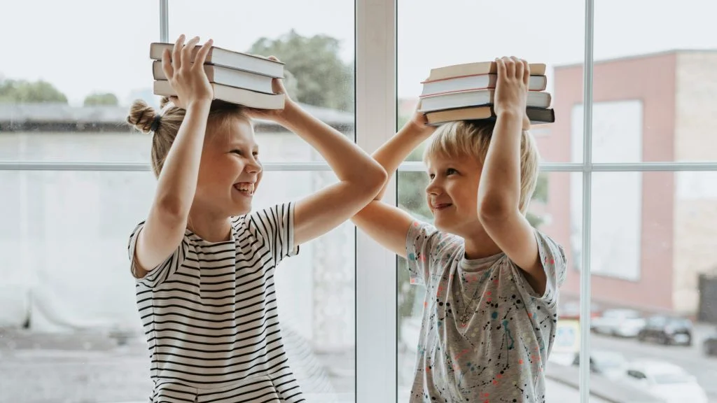 Two children balance books on their heads while smiling at each other by a large window.