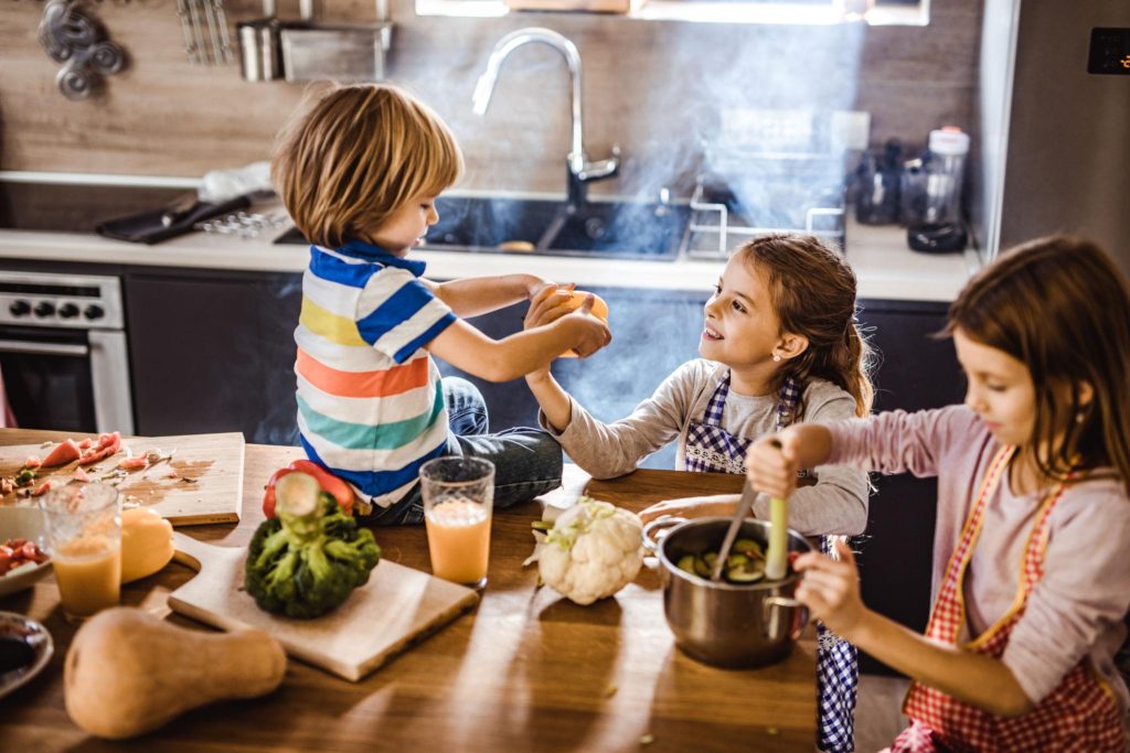 Children cooking together at a kitchen island, passing ingredients and smiling as they prep vegetables and snacks.