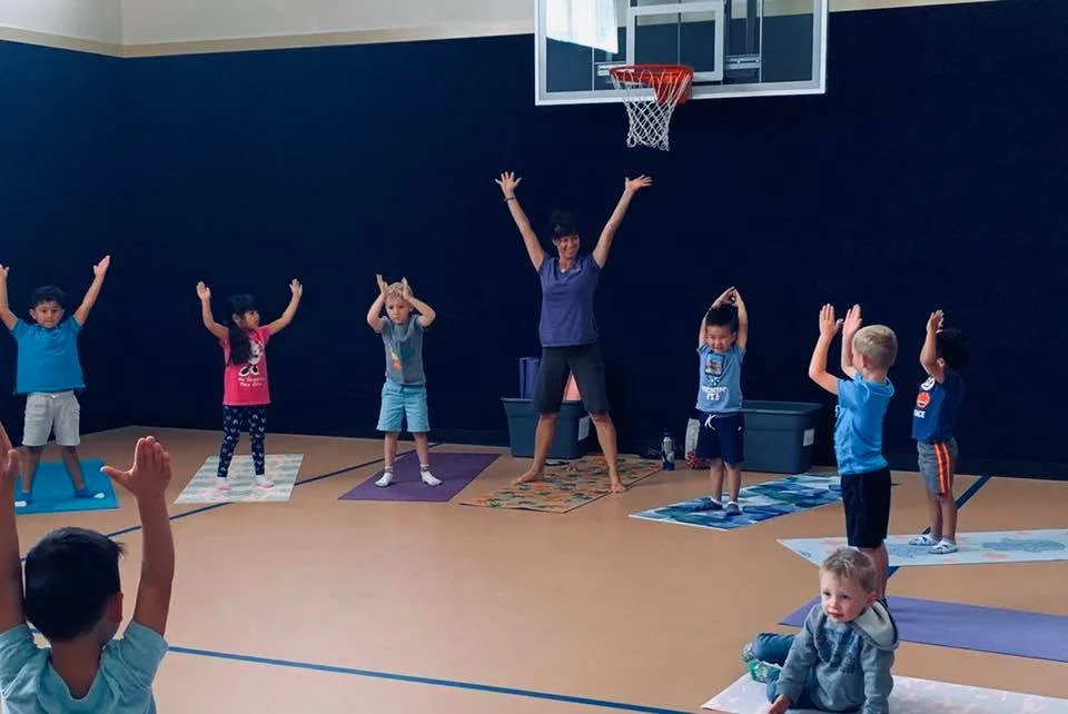 Instructor leading a children's yoga class in a gym, all kids on colorful mats with arms raised overhead.
