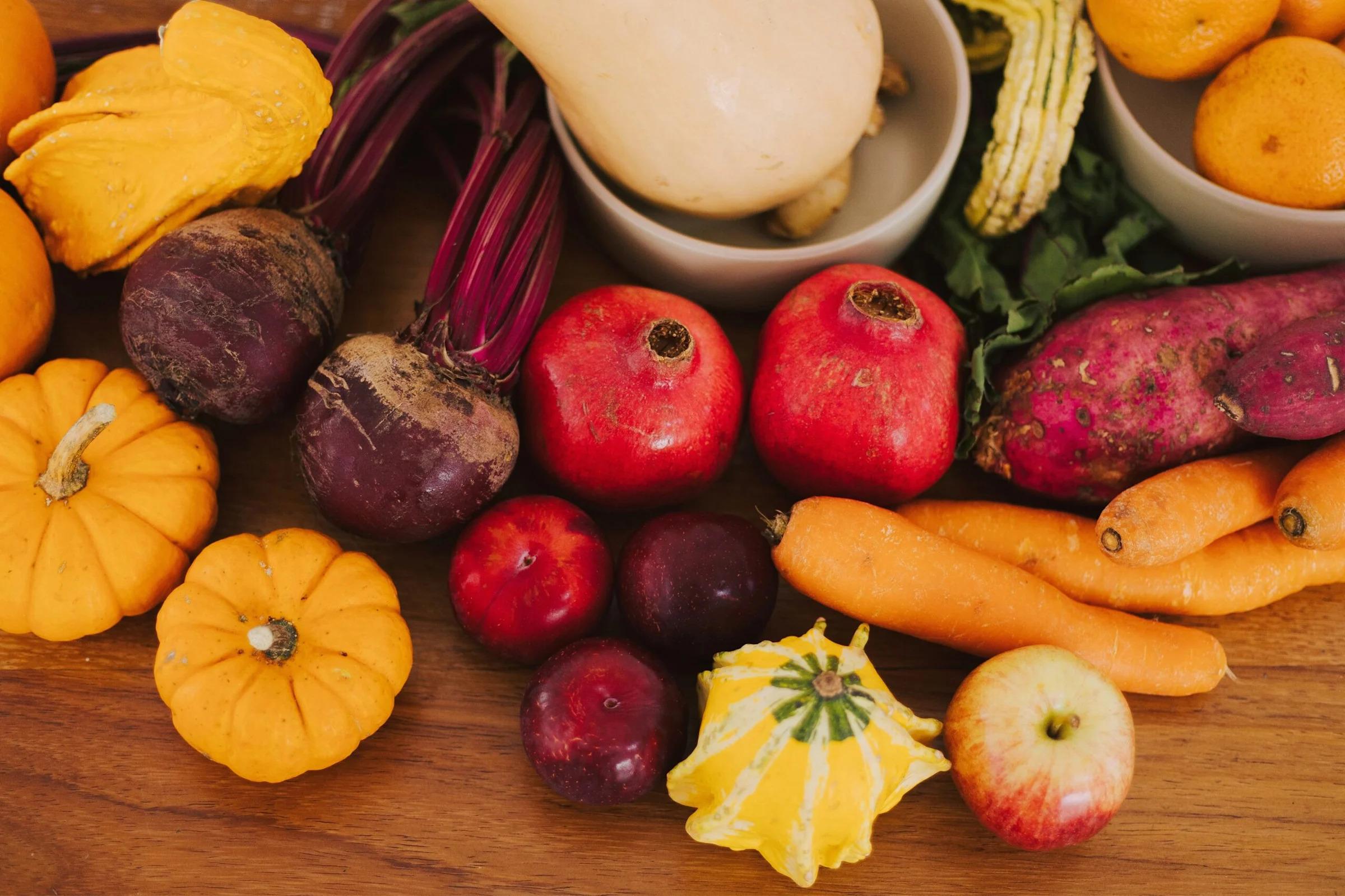 Assorted autumn produce on a wooden table, including pumpkins, apples, beets, carrots, and gourds with warm tones.