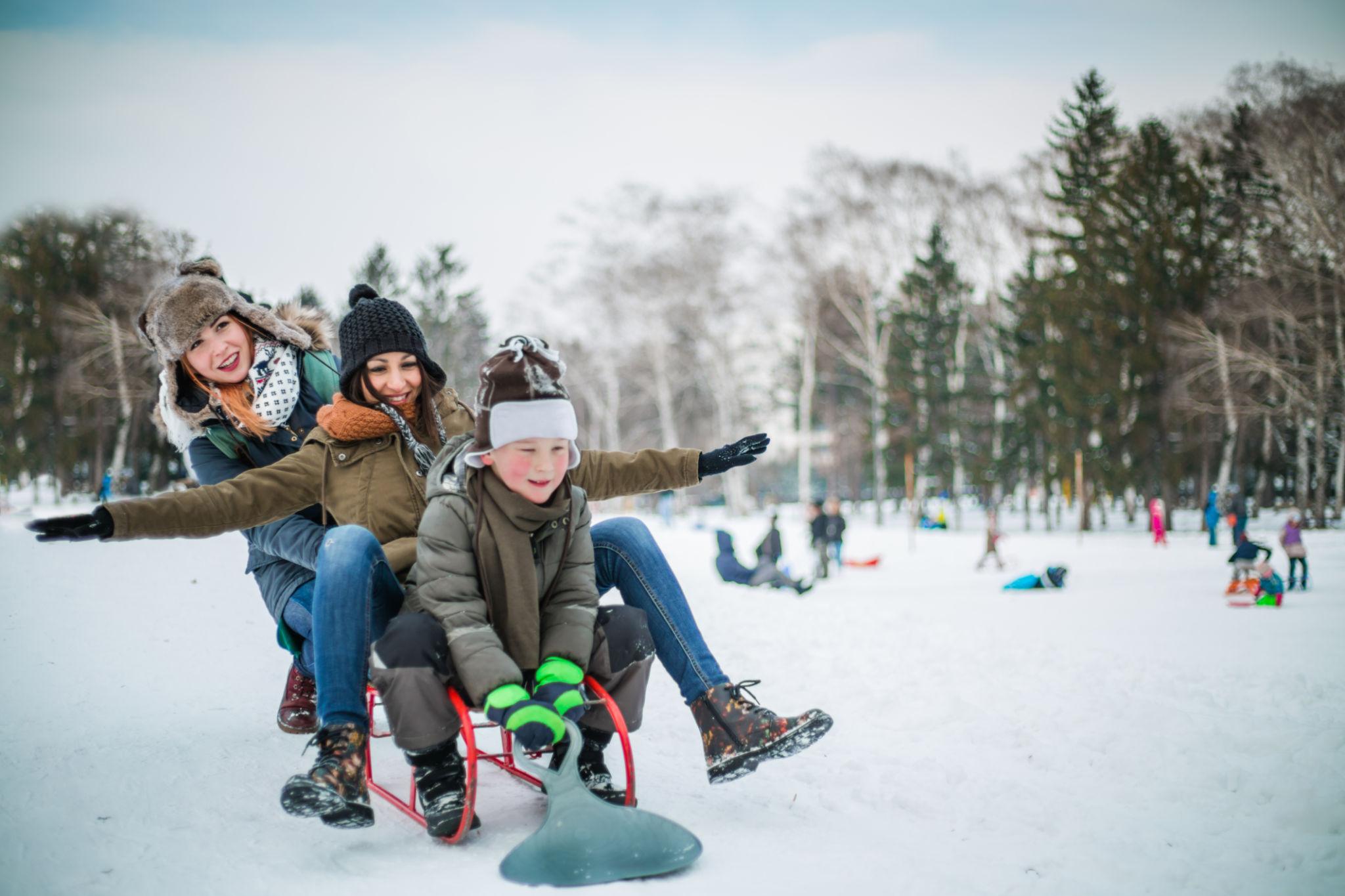 Three people on a red sled zoom across a snowy park, smiling in winter coats and hats.